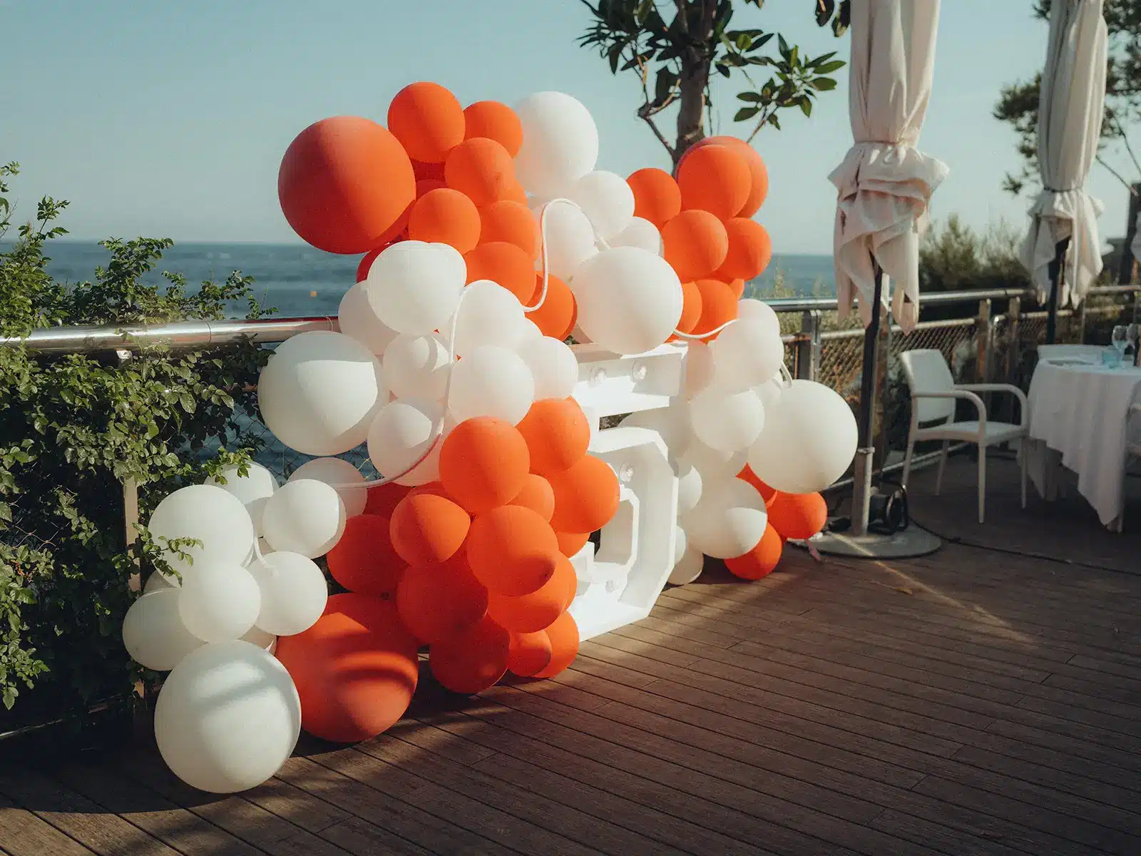 A festive outdoor setting with red and white balloons forms a decorative arch. Ocean view in the background, surrounded by greenery.