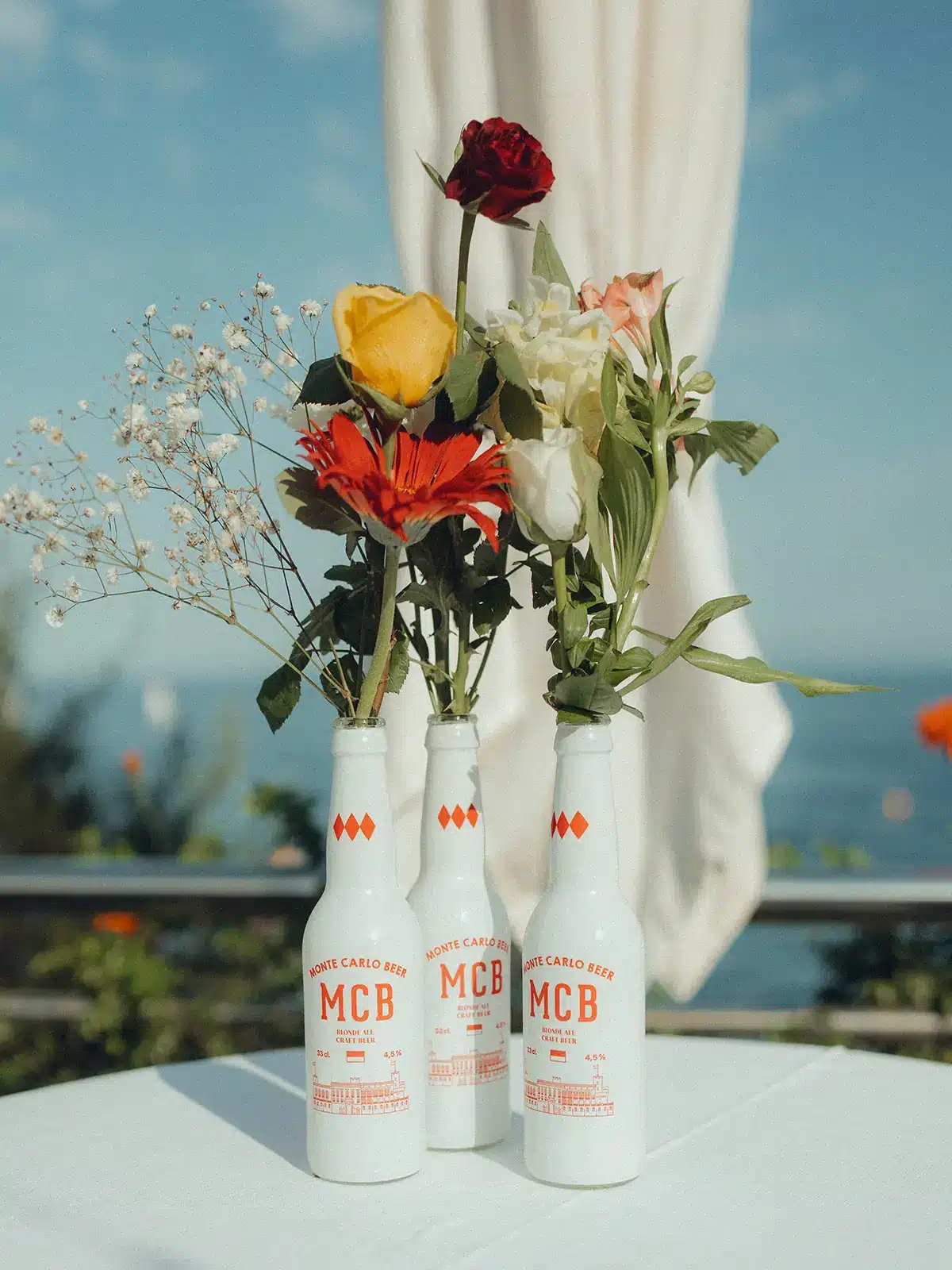 Three Monte Carlo Beer bottles with colorful flowers inside, on a table. Bright sky and railing in the background, suggesting an outdoor setting.