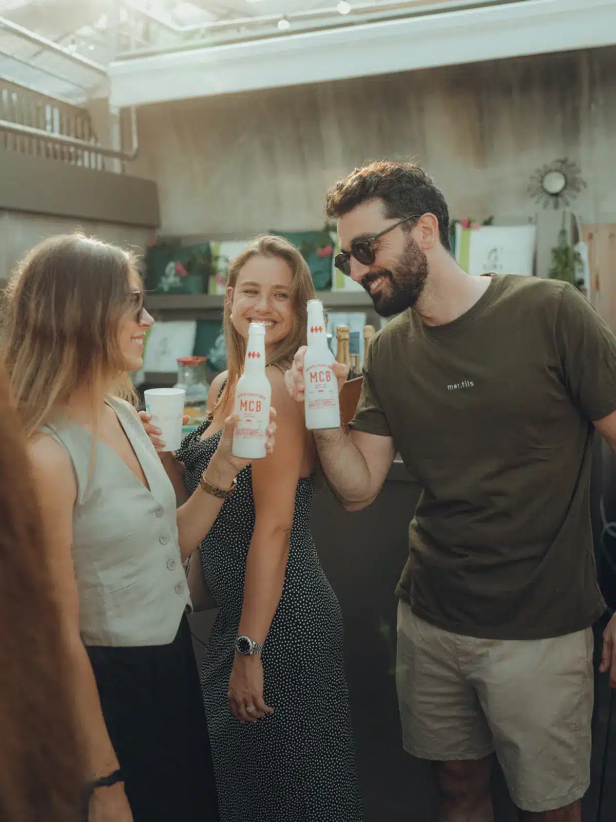 Three people smiling and toasting with bottles in a casual, well-lit outdoor setting, surrounded by plants and decor.