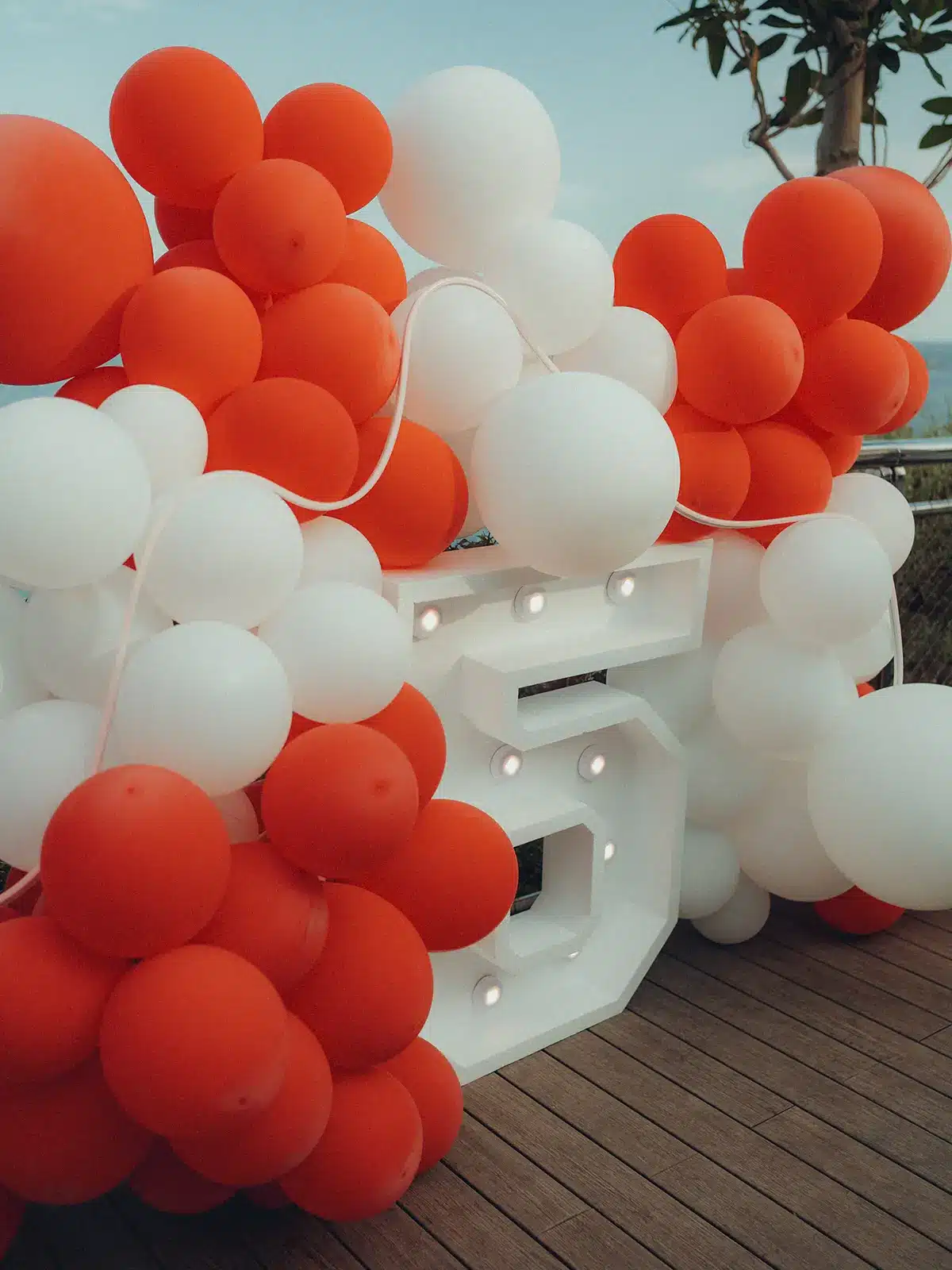 Orange and white balloons surround a large illuminated number 5 on a wooden deck, with trees and railing in the background.