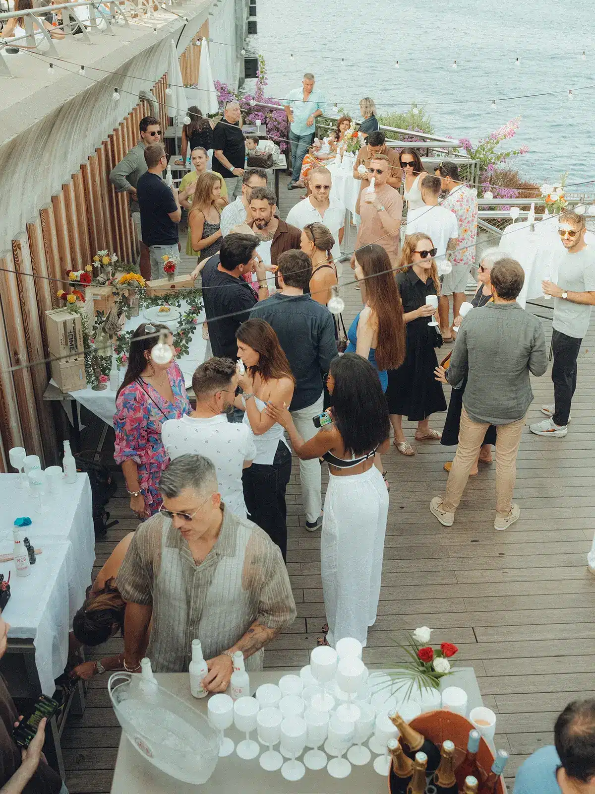 A gathering of people on a wooden deck by the water, enjoying drinks and conversation under string lights, surrounded by floral decor.