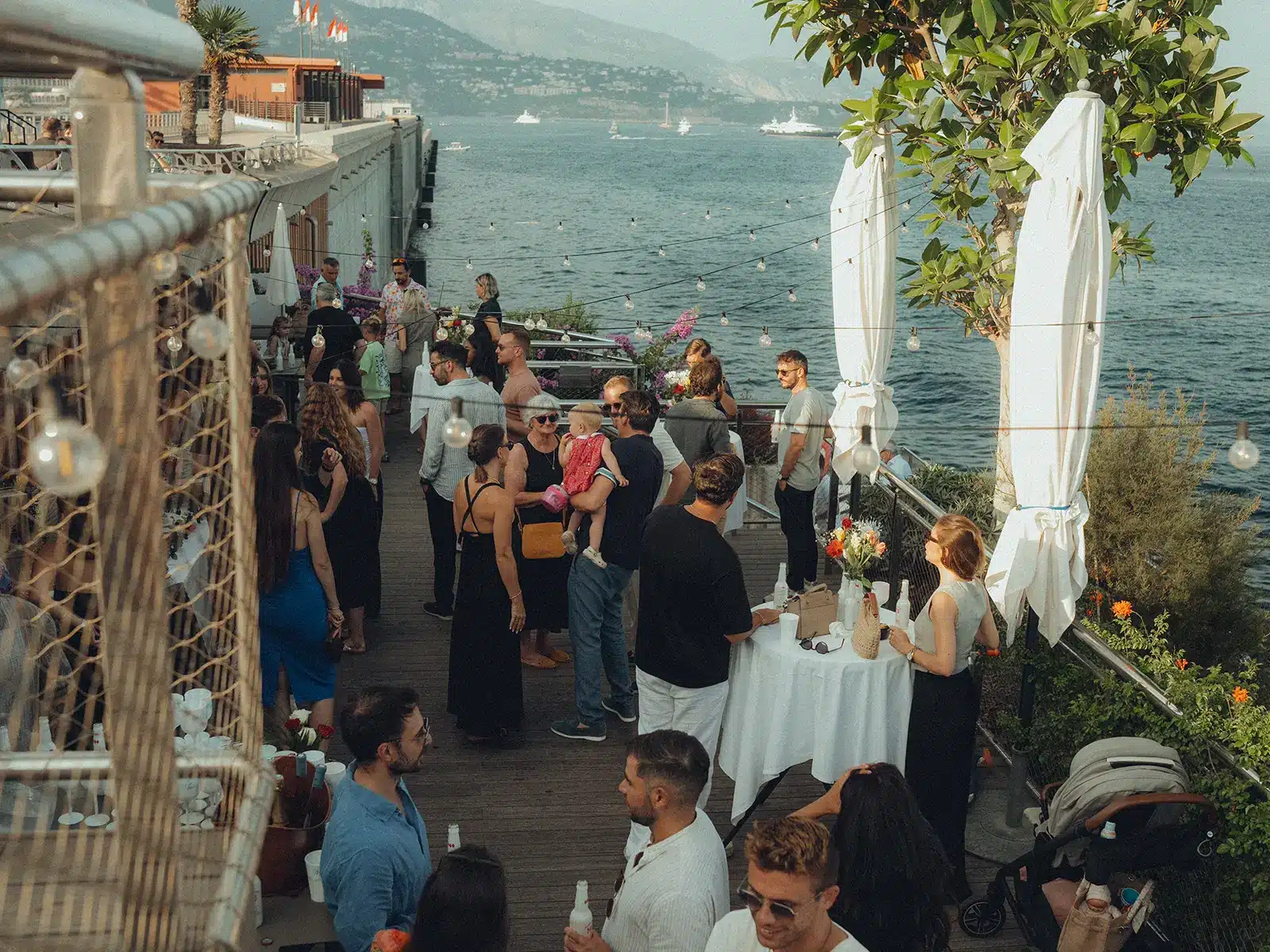 People gather on a deck overlooking a scenic waterfront, socializing amidst decorations and string lights, set against a backdrop of distant hills and boats.