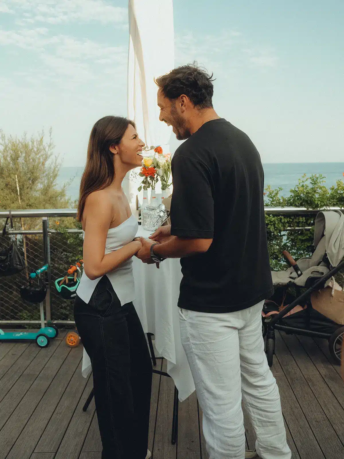 Two people stand closely on a deck overlooking the ocean, holding hands and smiling, with a stroller and greenery in the background.