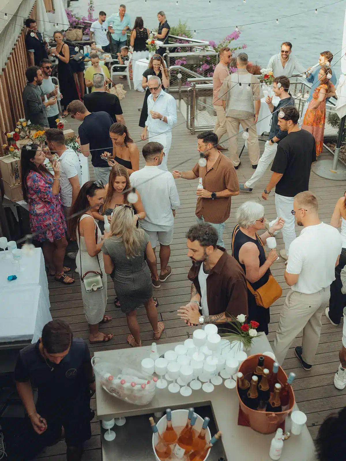 A group of people socializing at an outdoor gathering near water, featuring a bar setup with drinks and decorative lighting overhead.