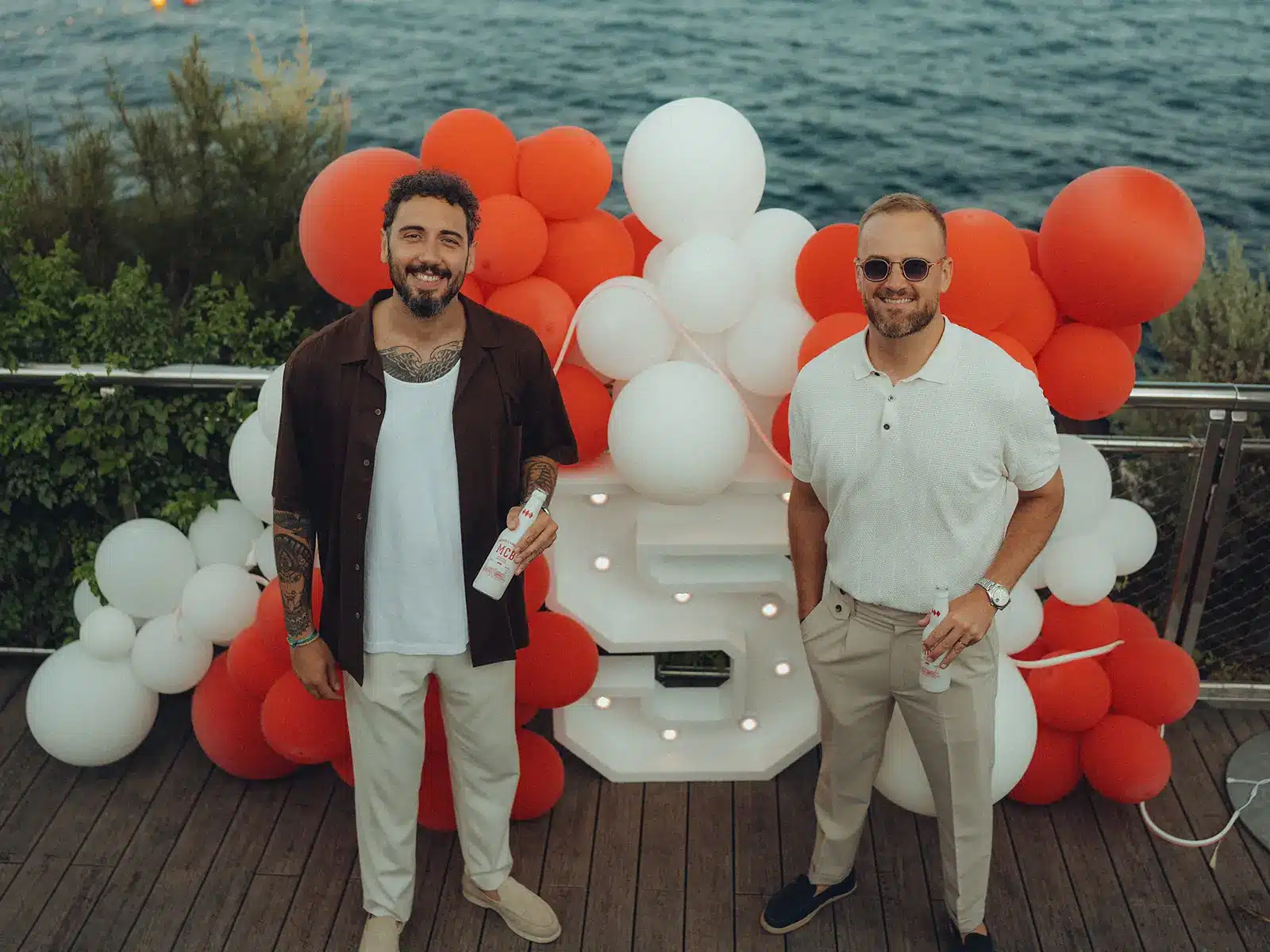 Projects 3 Two people smiling by a waterfront, surrounded by red and white balloons, on a wooden deck.
