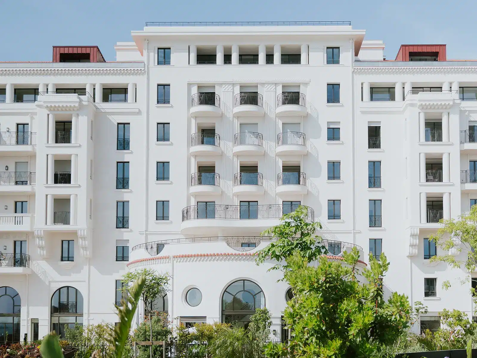 Projects 1 Elegant multi-story white building with balconies and arched windows, surrounded by greenery, under a clear blue sky. No identifiable landmarks or people visible.