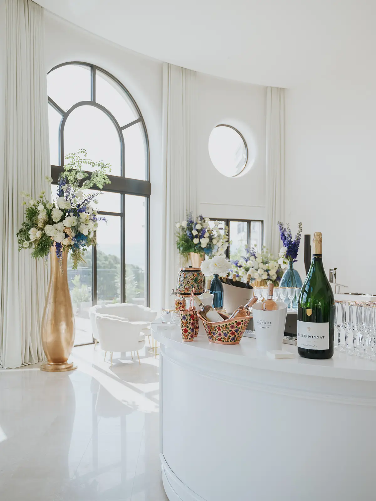 Elegant reception area with large arched windows, floral arrangements, and a white counter displaying wine bottles and glasses. Sunlight floods the bright interior.
