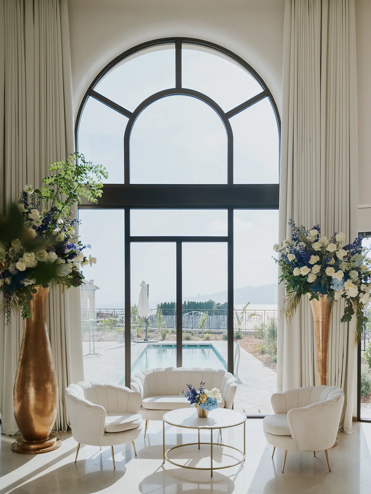 Elegant interior with large arched window, overlooking a pool. White chairs, gold table, and floral arrangements create a luxurious atmosphere.
