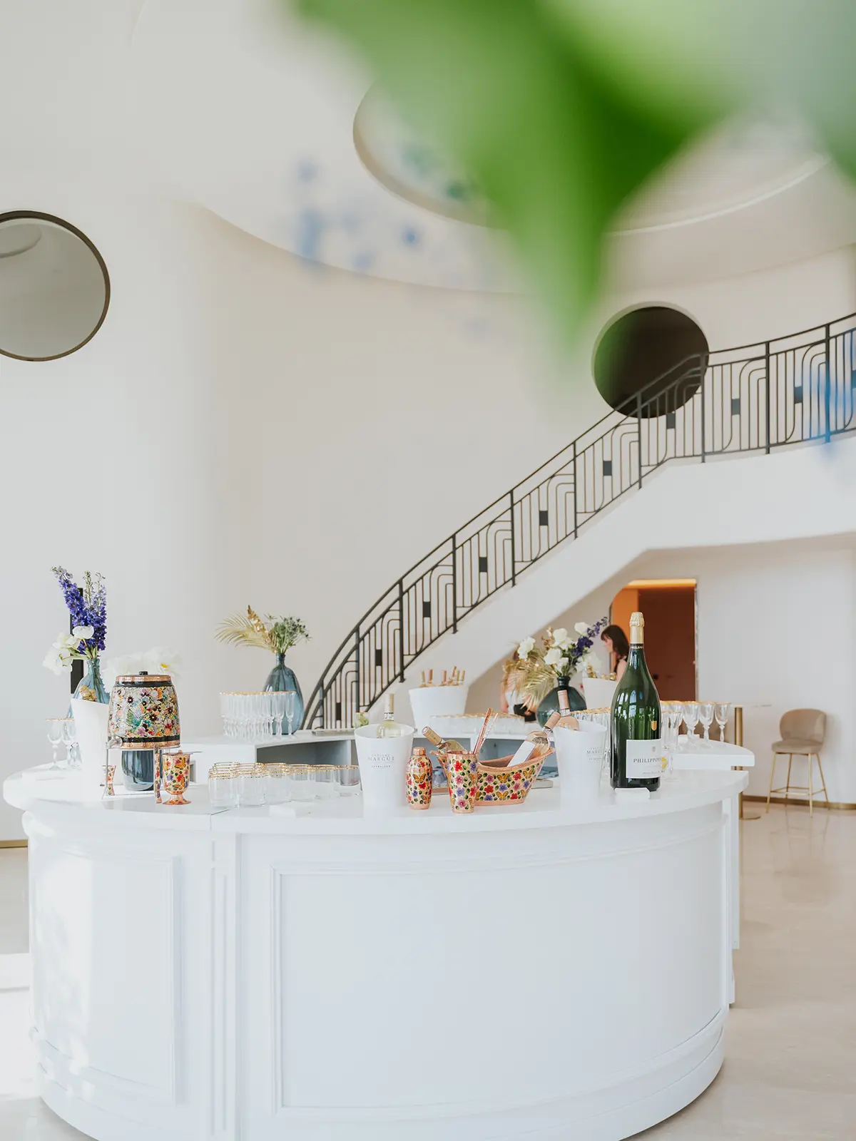 Elegant white room with a curved staircase, floral arrangements, large wine bottle, and drinks on a bar. A person stands in the background.