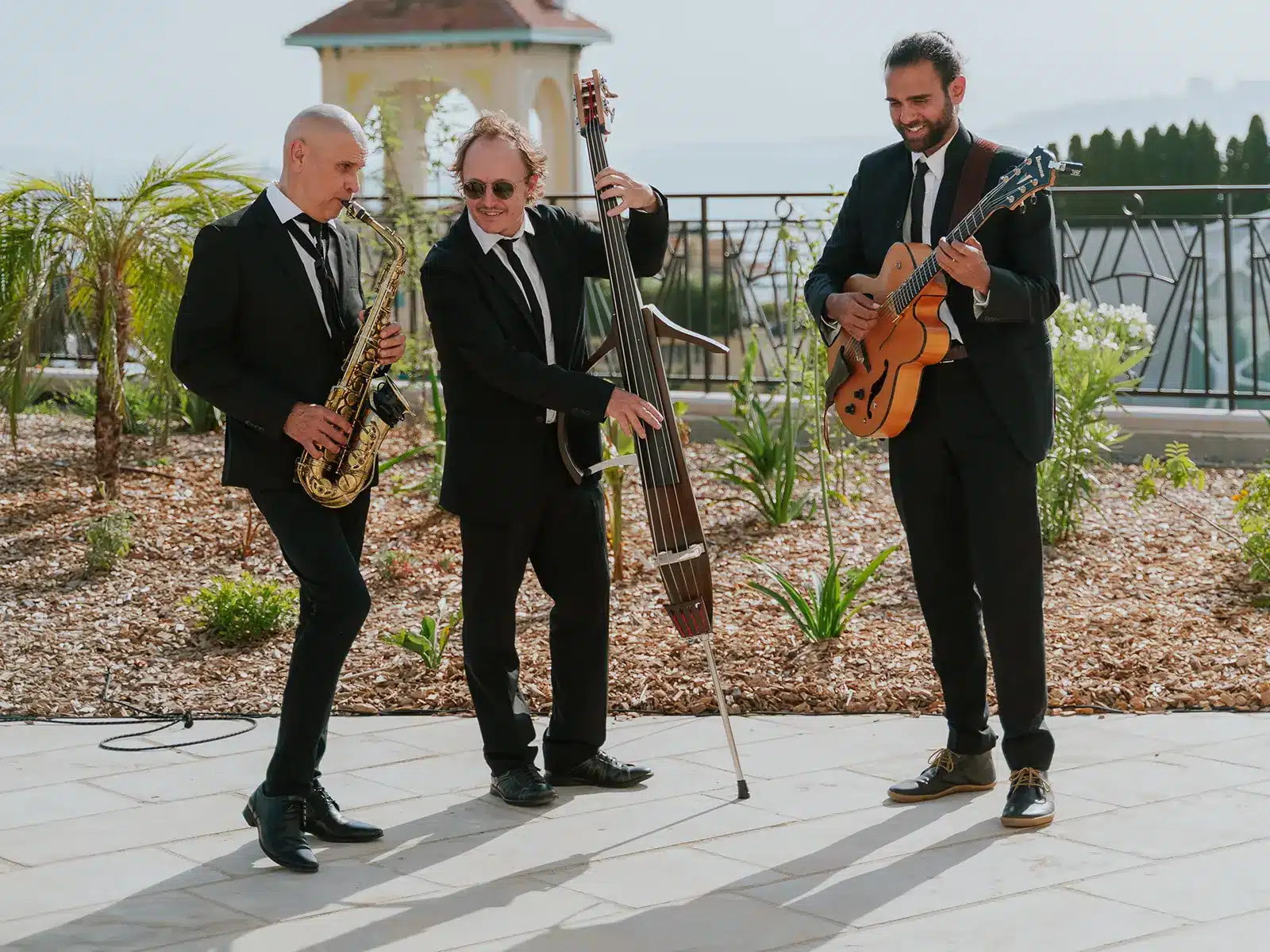 Three people in suits play jazz instruments outdoors near a garden, with a scenic view and a gazebo structure in the background.