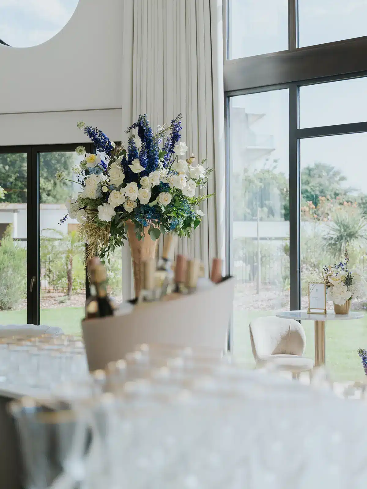 A modern, elegant room with large windows, floral arrangements, and a table set for an event. Bright, airy atmosphere with garden views.