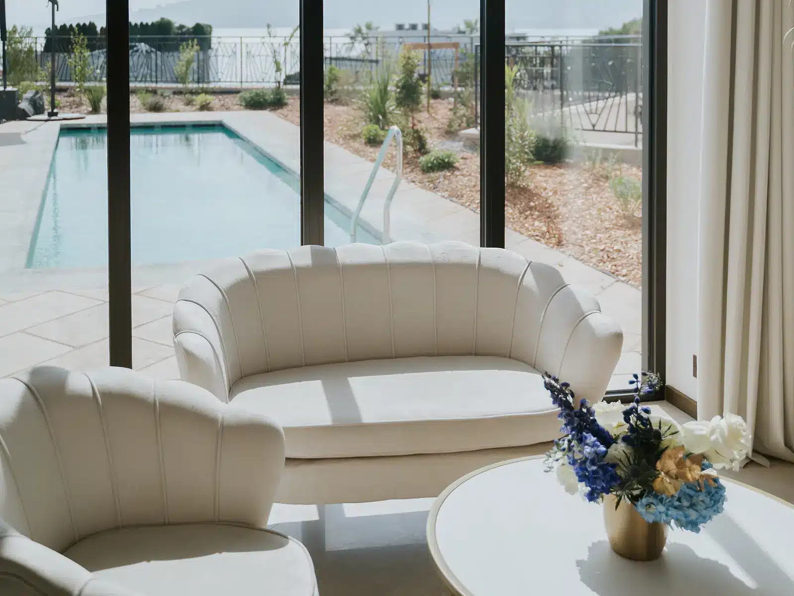 Cozy room with white sofas and flowers overlooking a swimming pool through large glass doors. Bright and airy interior with outdoor view.