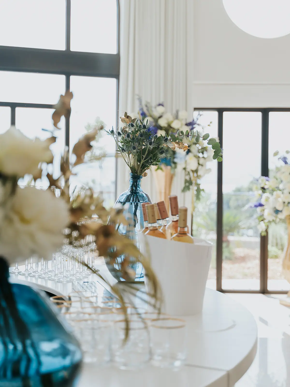Elegant setup with floral arrangements, wine bottles, and glassware on a table by large windows. Bright, natural light illuminates the scene.