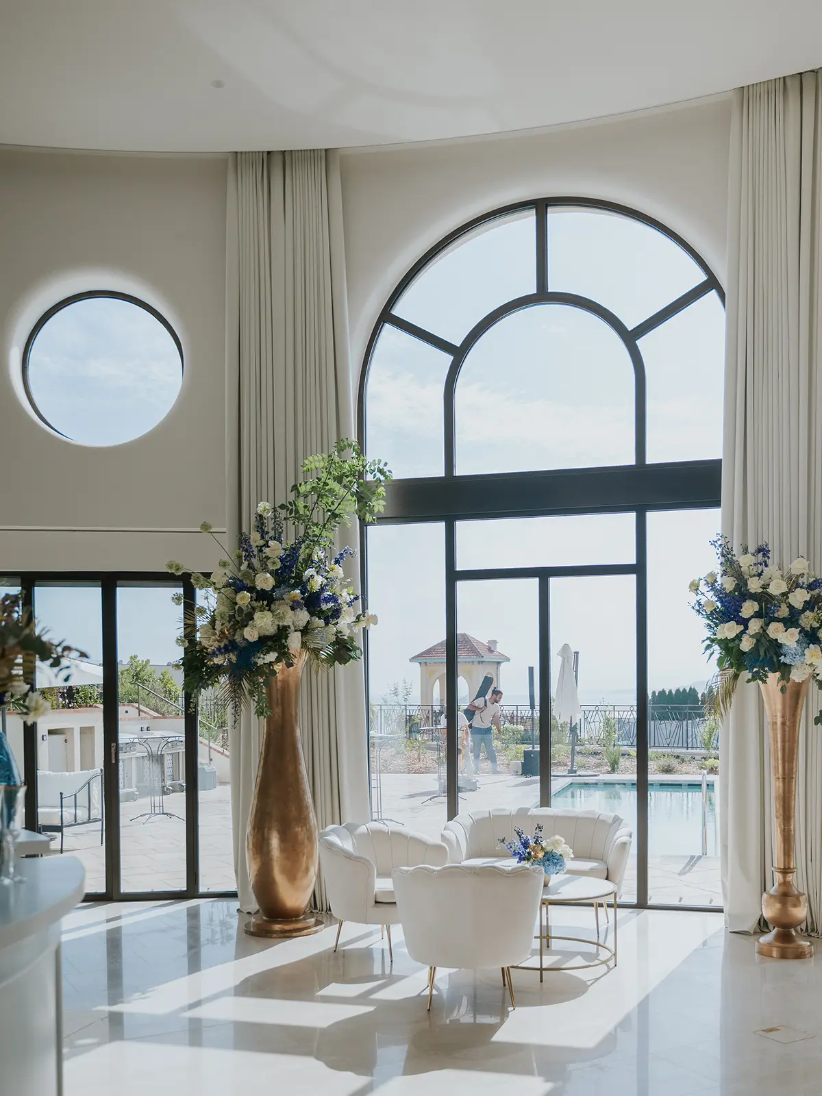 Elegant interior with large arched windows, round tables, and white chairs. Tall floral arrangements accent the space. Person seen by the pool outside.