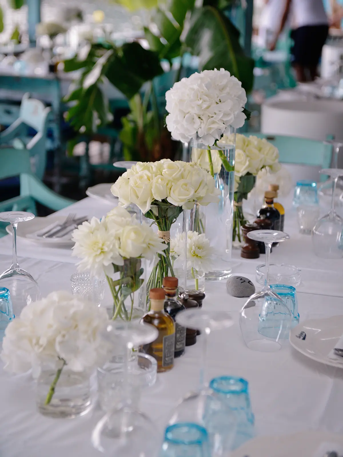 A table set for dining with white flowers, wine glasses, and condiments. The background shows a person and tropical plants.