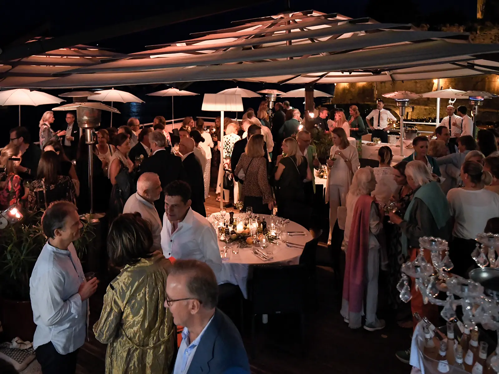 A large group of people socializing at a nighttime outdoor event with decorated tables and umbrellas. Stone wall visible in background.