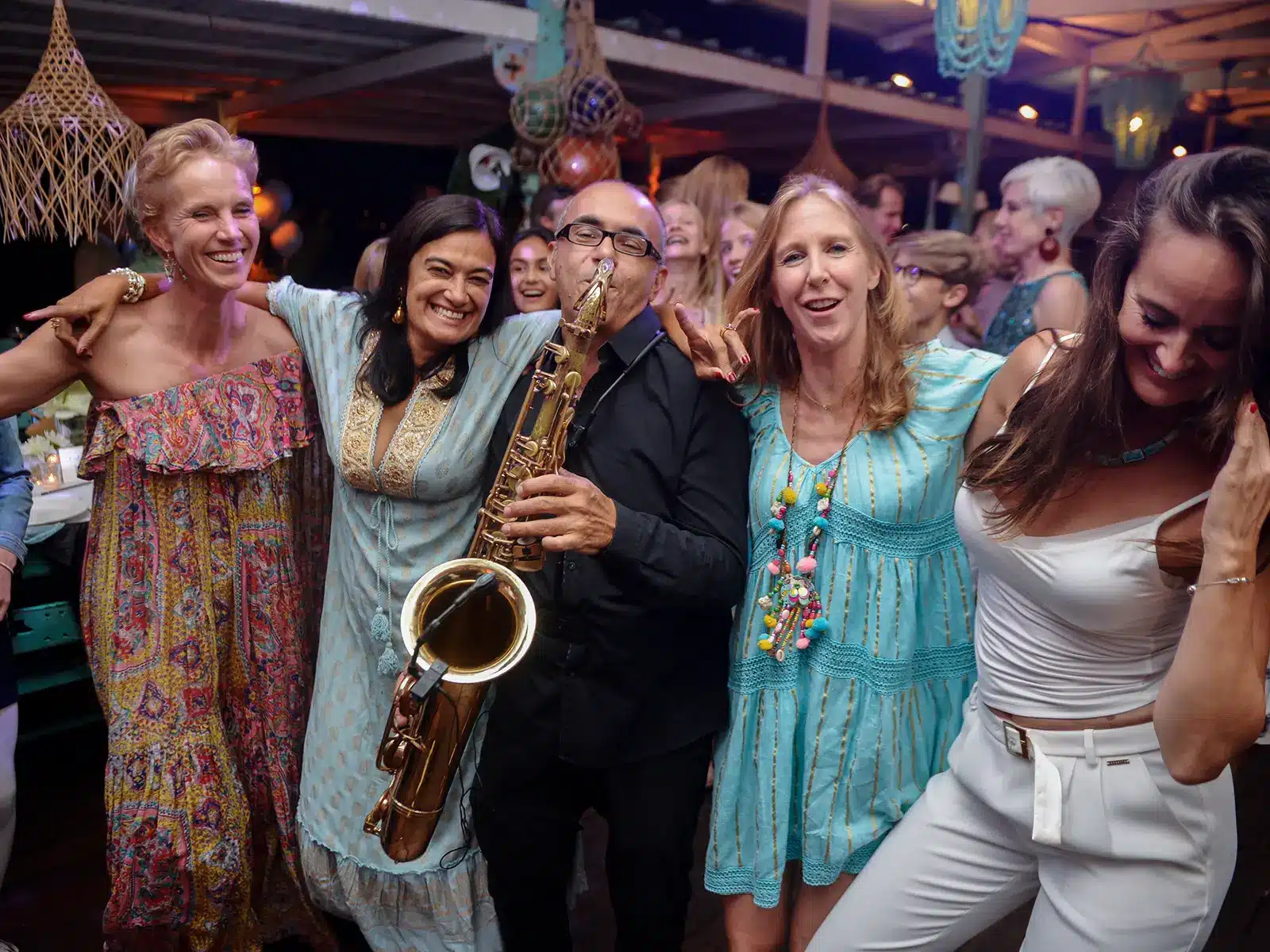 A lively gathering features a saxophonist and happy people dancing closely, creating a joyful, festive atmosphere under warm ambient lighting.