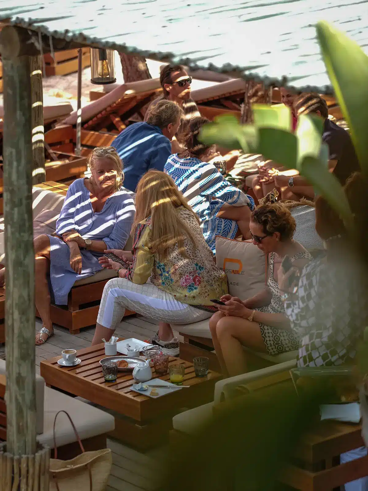 A group of people relax and socialize on outdoor sofas with drinks and snacks, under a shaded area, surrounded by greenery.