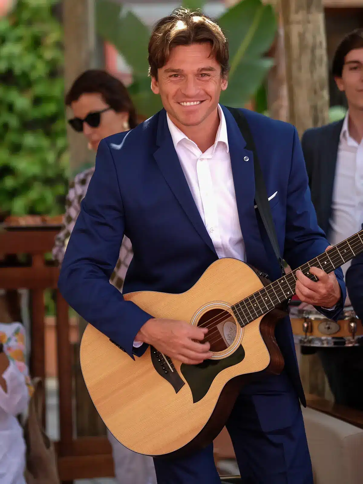 A person in a blue suit smiles while playing guitar, surrounded by musicians. Green foliage is visible in the background.
