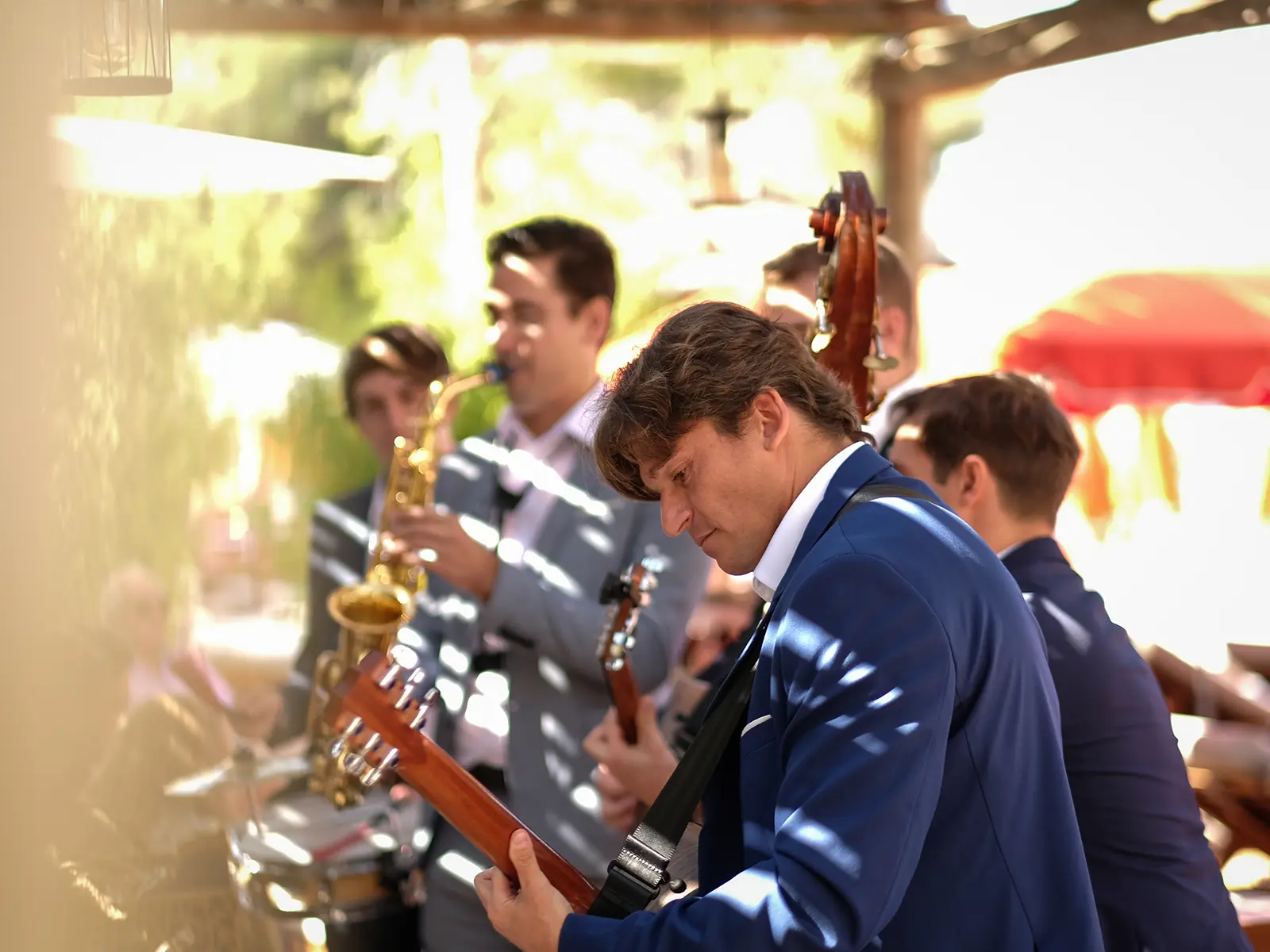Musicians in blue suits perform outdoors, playing guitar and saxophone. Sunlight filters through, creating a lively, warm atmosphere. No recognizable landmarks are visible.