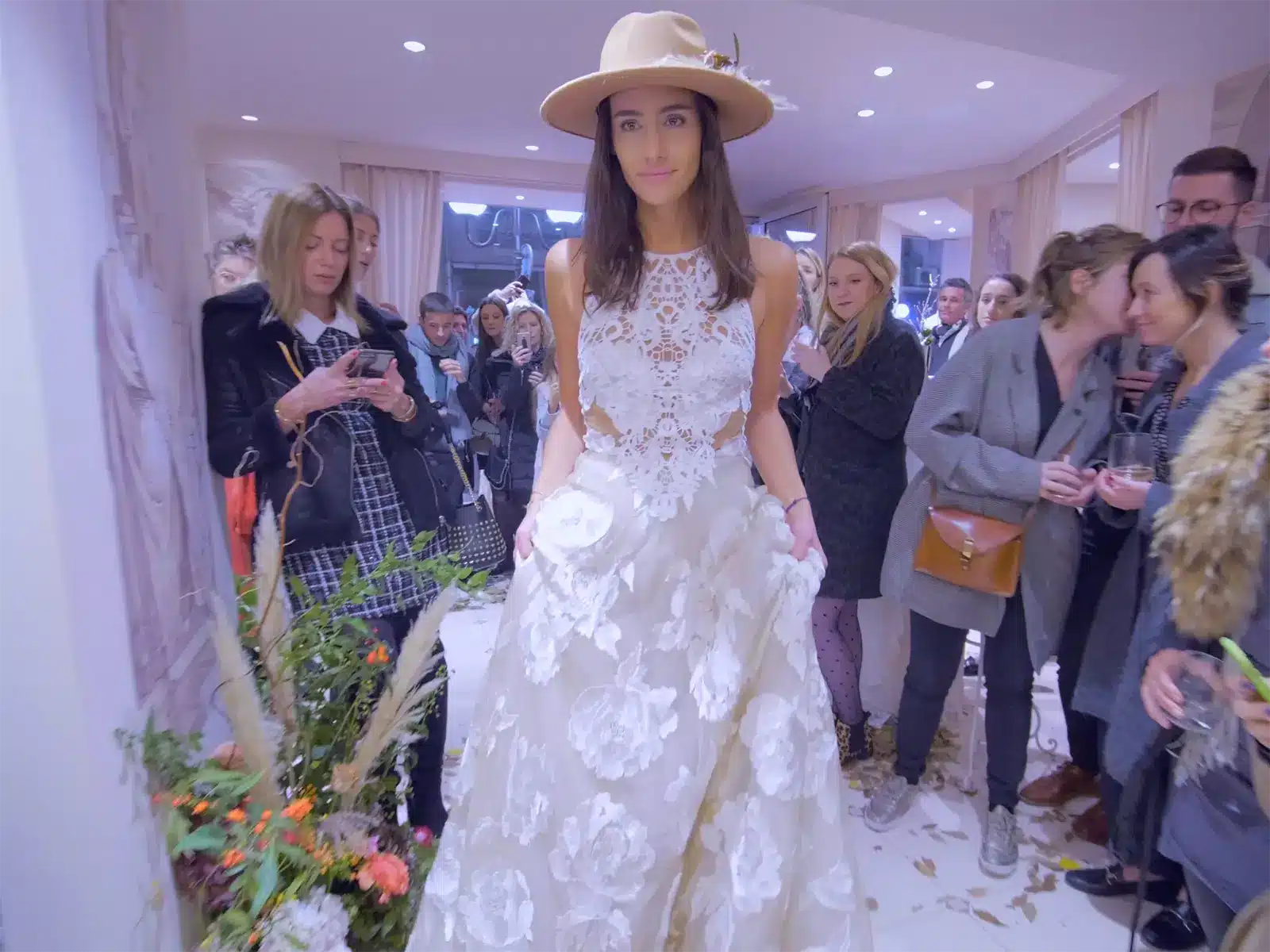 Projects 4 A person in a floral dress and hat poses in a crowded room. People gather around, some using phones.